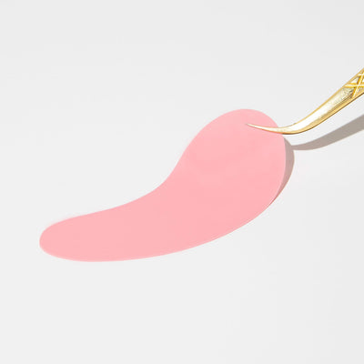 Curacoro gold tweezers holding a flat pink silicone lash pad on a white background.