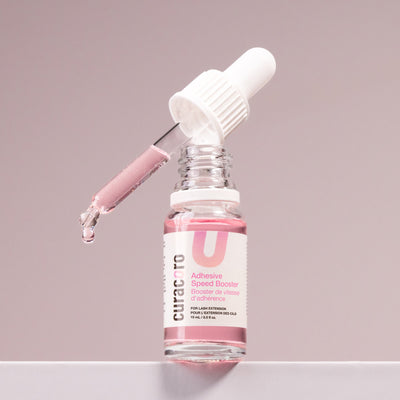 Curacoro Adhesive Speed Booster in a clear bottle with a dropper, filled with pink liquid for lash extensions.