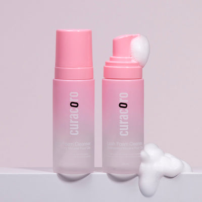 Two pink bottles with foam spilling from one, set against a soft white and pink background.