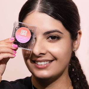 A smiling woman with braided hair holding a Curacoro Promade Loose Fans case near her eye against a soft pink background.
