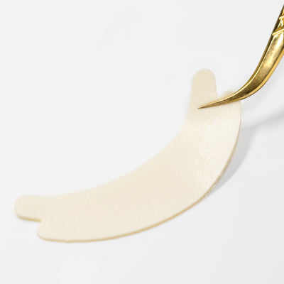 A close-up of a single hypoallergenic foam under-eye pad being lifted with gold tweezers on a white background.