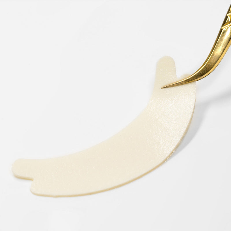 A close-up of a single hypoallergenic foam under-eye pad being lifted with gold tweezers on a white background.