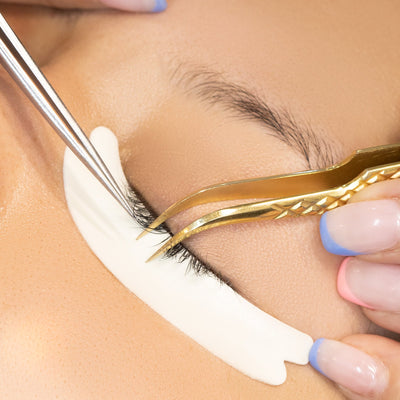 A close-up view of a model's closed eye with a hypoallergenic foam under-eye pad applied, while gold tweezers are used for precise lash application, showcasing professional lash extension techniques.