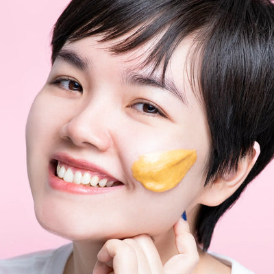 A smiling woman applying a gold hydro gel mask to her cheek against a pink background.