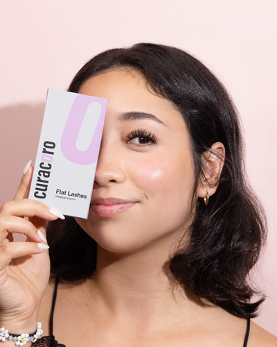 A woman with a radiant complexion holds a Curacoro Flat Lashes product box near her face, with a soft pink background.