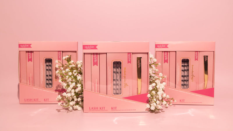 Three pink lash kit boxes arranged in a row, featuring DIY lash extension sets from Quickie by LLBA, with delicate white flowers in the foreground against a soft pink background.