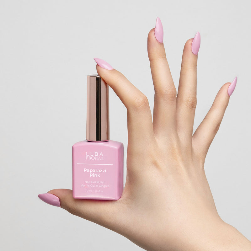 Hand holding a bottle of LLBA Pronail Paparazzi Pink Nail Gel Polish, featuring a light pink shade, with matching nail color on the hand, against a light background.