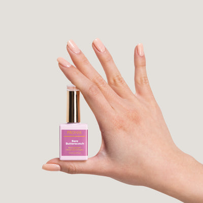 A hand with glittery Bare Butterscotch nails holding a nail polish bottle with a gold cap against a light beige background.