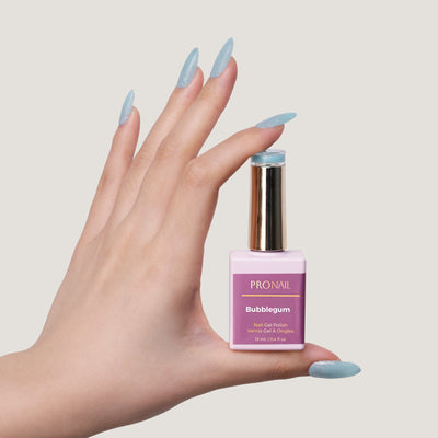 A hand with glittery Bubblegum nails holding a nail polish bottle with a gold cap against a light beige background.