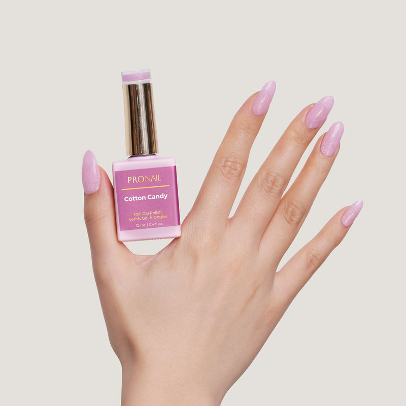 A hand with glittery Cotton Candy nails holding a nail polish bottle with a gold cap against a light beige background.