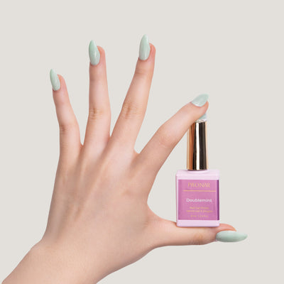 A hand with glittery Doublemint nails holding a nail polish bottle with a gold cap against a light beige background.