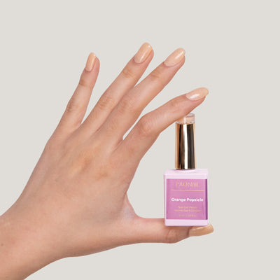 A hand with glittery Orange Popsicle nails holding a nail polish bottle with a gold cap against a light beige background.