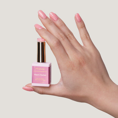 A hand with glittery Peach Parfait nails holding a nail polish bottle with a gold cap against a light beige background.