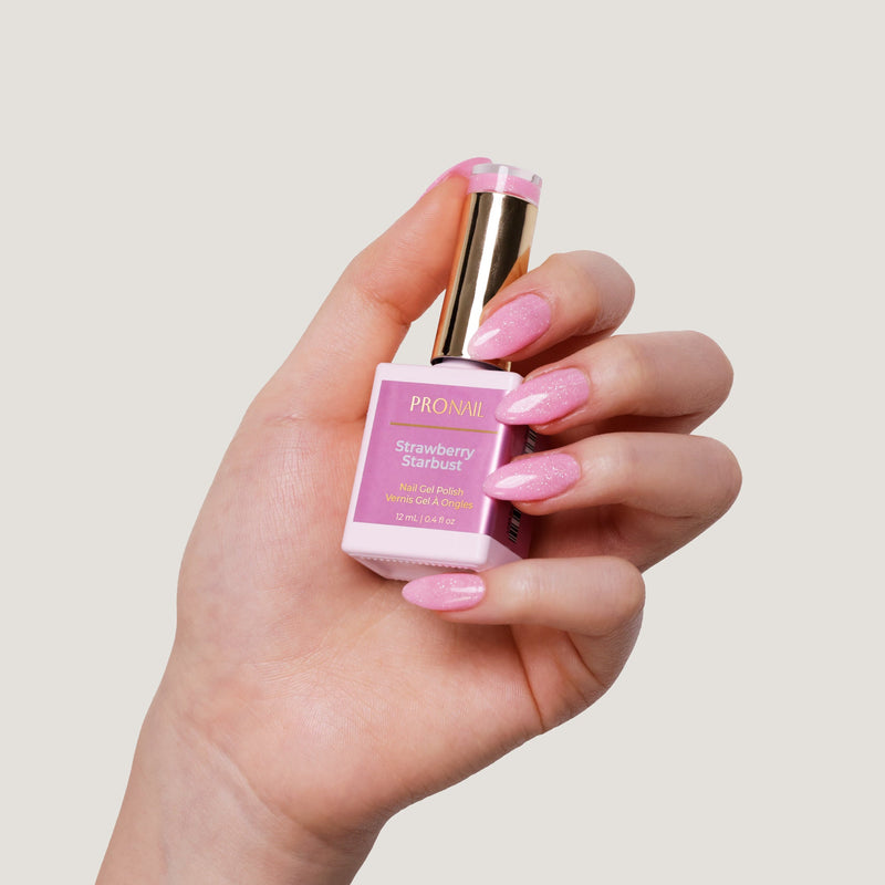 A hand with glittery Strawberry Starbust nails holding a nail polish bottle with a gold cap against a light beige background.