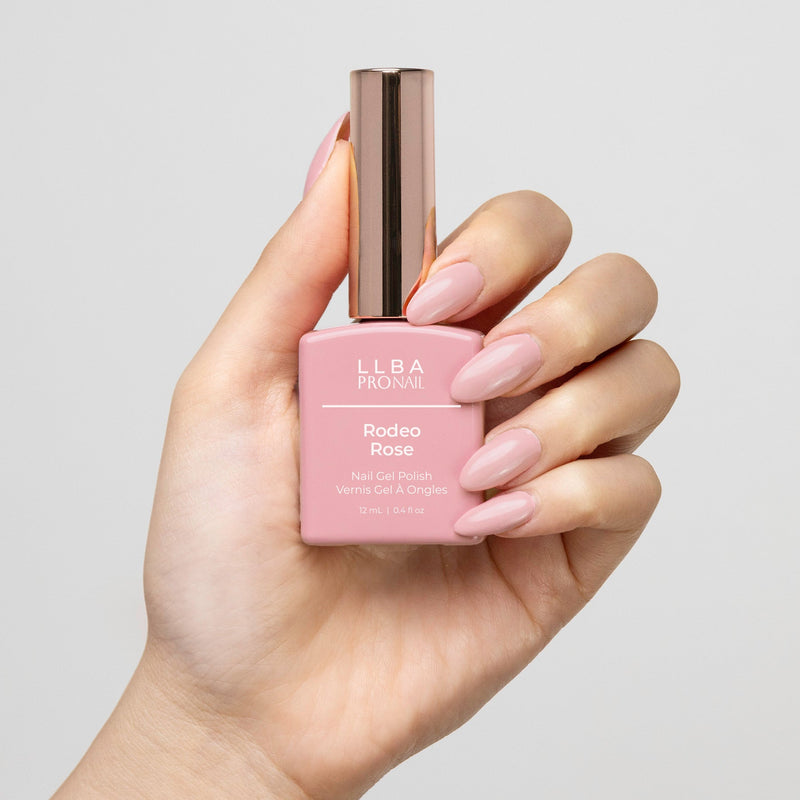 Hand holding a bottle of LLBA ProNail Rodeo Rose nail gel polish, featuring light pink nails matching the color of the polish. The bottle has a gold cap with the product name visible on the front.