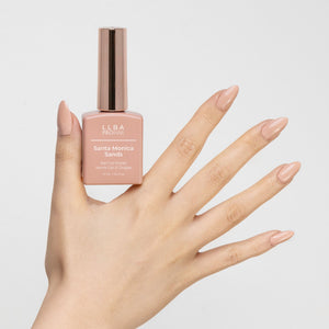 Hand holding a bottle of LLBA ProNail Santa Monica Sands nail gel polish, featuring nails painted in a beige nude shade that matches the polish color. The bottle has a gold cap and the product name is visible on the front.