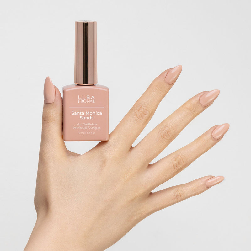 Hand holding a bottle of LLBA ProNail Santa Monica Sands nail gel polish, featuring nails painted in a beige nude shade that matches the polish color. The bottle has a gold cap and the product name is visible on the front.