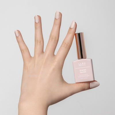 Hand holding a bottle of LLBA ProNail Scarlet Secret nail gel polish, with nails painted in a matching soft beige nude shade. The bottle features a gold cap and the product name is clearly visible on the front.