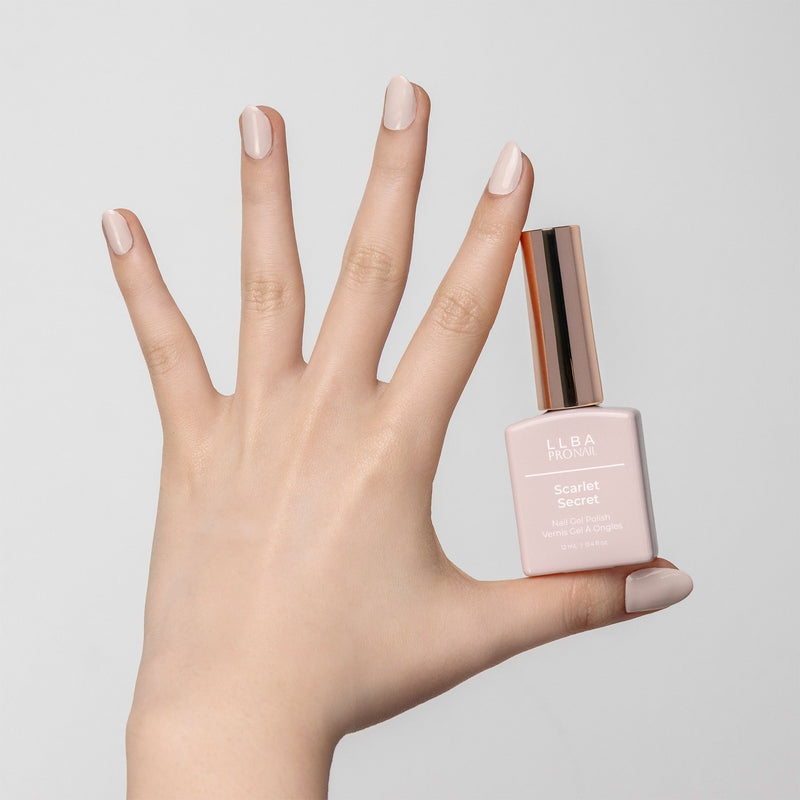 Hand holding a bottle of LLBA ProNail Scarlet Secret nail gel polish, with nails painted in a matching soft beige nude shade. The bottle features a gold cap and the product name is clearly visible on the front.