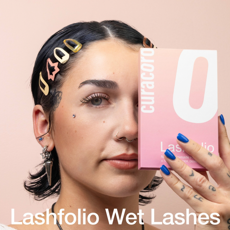 Model with short dark hair, colorful hairpins, and blue nails holding the Curacoro Lashfolio Wet Lashes box against a peach background.