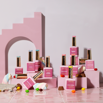 A playful arrangement of nail polish bottles with gold caps surrounded by colorful candies and marshmallows on a pink-tiled surface.