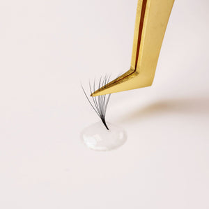 Gold tweezer holding lash extension dipped in clear glue on white background.